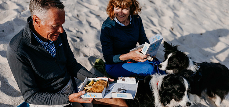 Couple eating Adams Fish and Chips takeaway on par beach with their dogs, St Martins, Isles of Scilly