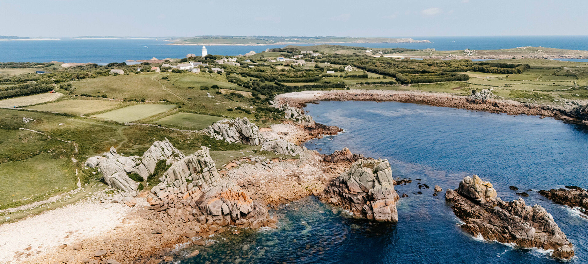 Drone view of St Agnes, Isles of Scilly, showing rugged coastline, turquoise waters, and St Agnes Lighthouse surrounded by green fields under a clear blue sky.